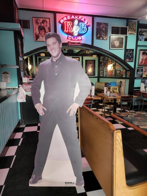 A life-sized cutout of Elvis Presley stands in a retro diner with colorful decor and a neon sign.