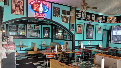 A retro diner interior with turquoise walls, checkered floor, and a neon "Breakfast Club" sign, adorned with vintage photos.