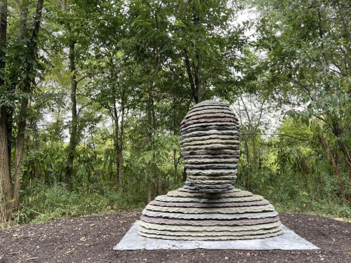 A layered stone sculpture of a head and shoulders, surrounded by lush greenery in a natural setting.