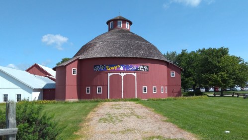 A red round barn with a sign for "Guys and Dolls" on display, surrounded by green grass and trees under a blue sky.