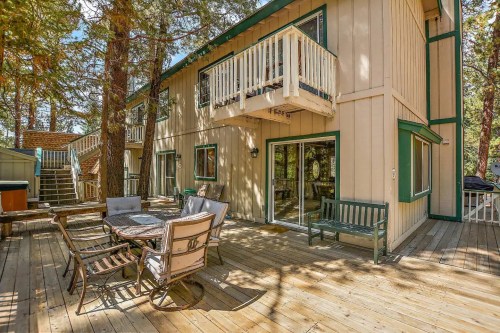 A wooden deck with outdoor seating, surrounded by trees, next to a two-story house with a balcony.