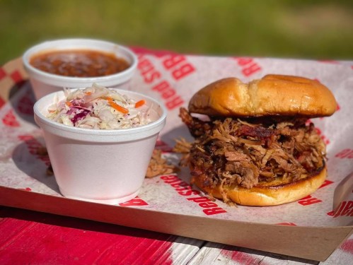A pulled pork sandwich on a bun, served with coleslaw and a cup of baked beans on a red and white tray.
