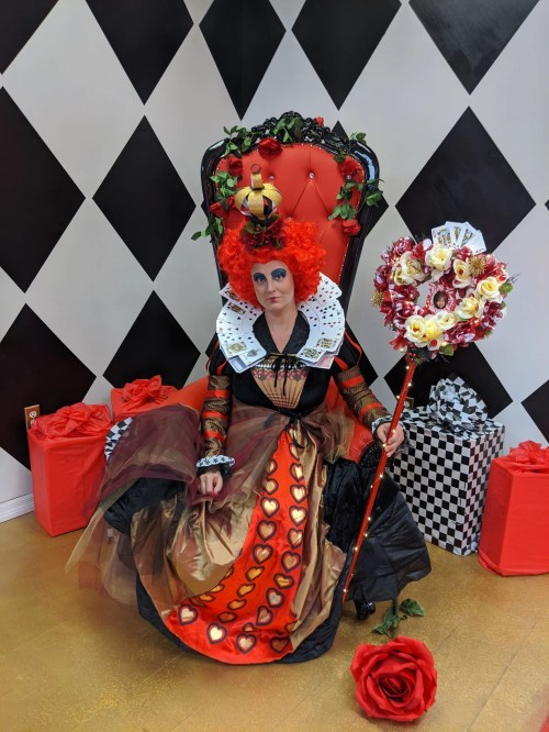 A woman dressed as the Queen of Hearts sits on a red throne, surrounded by gifts and holding a decorated staff.