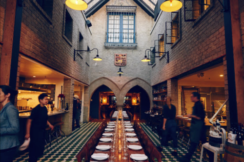 A spacious restaurant with a long table, high ceilings, and a mix of modern and rustic decor. Staff are serving guests.