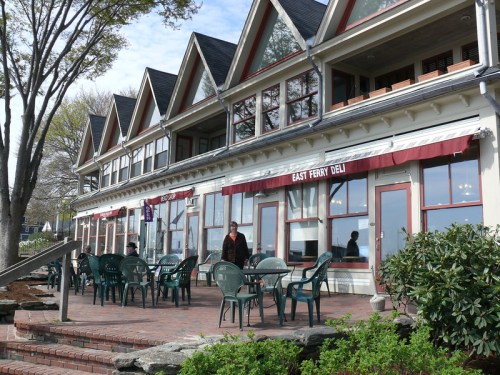 A waterfront deli with outdoor seating, featuring a brick patio and large windows under a blue sky.