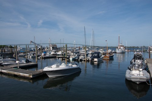 A busy marina with various boats docked, under a clear blue sky and a bridge in the background.