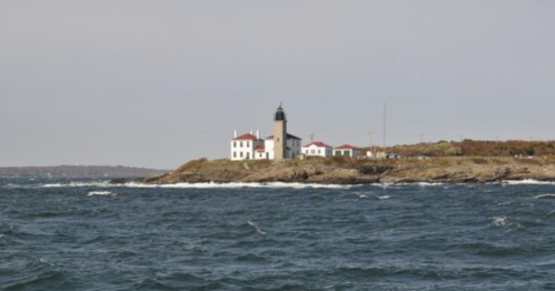 A white lighthouse with a black tower stands on a rocky shore, surrounded by choppy blue waters under a gray sky.
