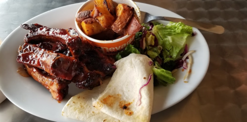 A plate with BBQ ribs, roasted potatoes, a side salad, and a tortilla on a metal table.