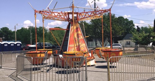 A colorful carnival ride with empty seats, surrounded by barriers and portable restrooms in a sunny outdoor setting.