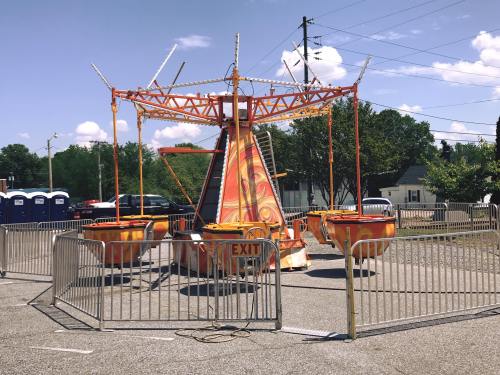 An empty carnival ride with orange and yellow colors, surrounded by a fence, under a clear blue sky.