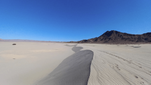 A vast desert landscape with a clear blue sky, featuring sandy dunes and a distant mountain range.