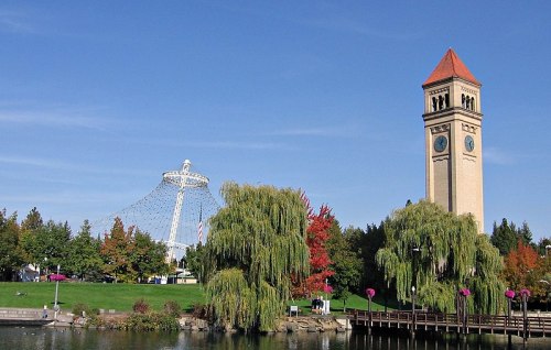 A scenic view of a park featuring a clock tower, a Ferris wheel, and colorful trees by a calm water body.