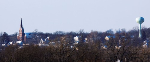 A winter landscape featuring a church steeple, water tower, and snow-covered houses against a clear sky.