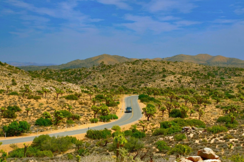 A winding road cuts through a desert landscape with mountains and sparse vegetation under a blue sky.