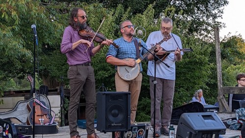 Three musicians perform outdoors, playing a violin, banjo, and mandolin, with a crowd and greenery in the background.