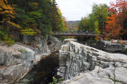 A wooden bridge spans a rocky gorge with autumn foliage in vibrant colors surrounding the scene.