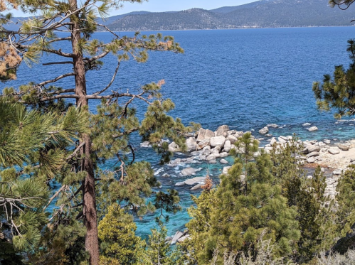 A scenic view of a blue lake surrounded by rocky shores and lush green trees under a clear sky.
