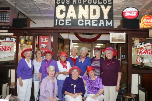 A group of elderly women in colorful outfits and hats pose in front of a vintage ice cream shop sign.