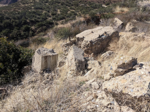 Rubble and large stone blocks scattered among dry grass and shrubs on a hillside.