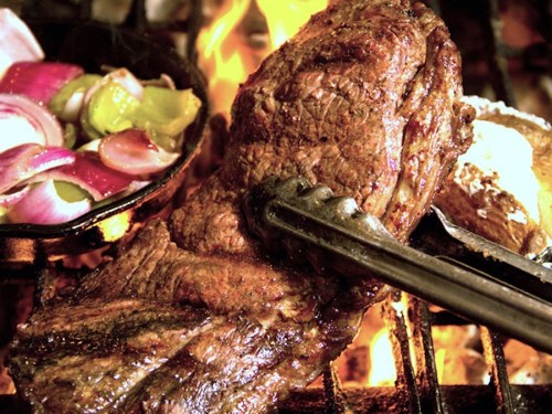 A close-up of grilled meat on a barbecue, with colorful vegetables in a skillet and flames in the background.