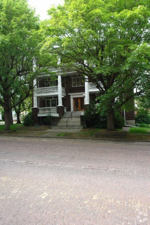 A large, two-story house with a porch, surrounded by green trees, located on a brick-paved street.