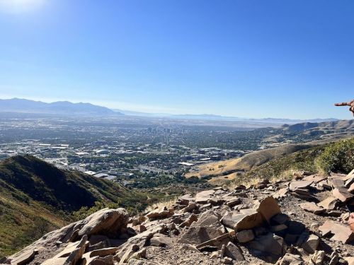 A panoramic view of a valley from a mountain, showcasing urban areas and distant mountains under a clear blue sky.