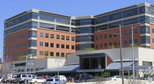 Modern multi-story building with large windows, featuring retail spaces and parking in the foreground. Clear blue sky.