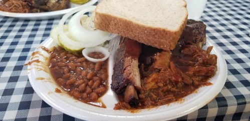 A plate of barbecue ribs with baked beans, onions, pickles, and a slice of bread on a checkered tablecloth.