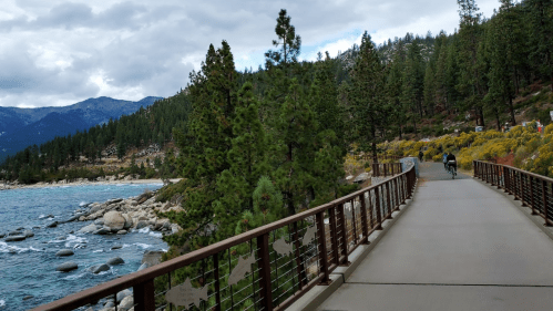 A scenic pathway along a lake, surrounded by trees and mountains, with two people walking in the distance.