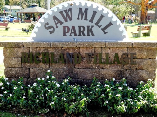 Sign for Saw Mill Park in Richland Village, featuring a decorative saw blade design and surrounded by flowers.