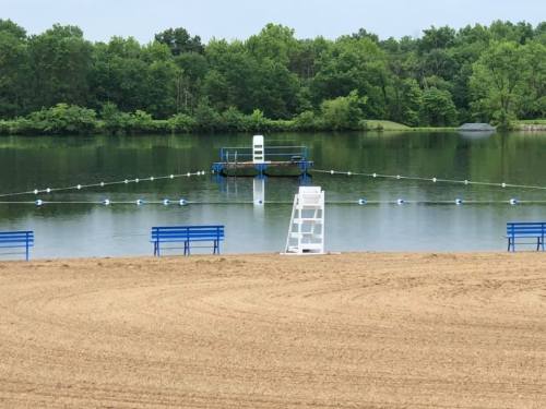 A calm lake with a floating platform, surrounded by blue benches and a lifeguard chair on the sandy shore.