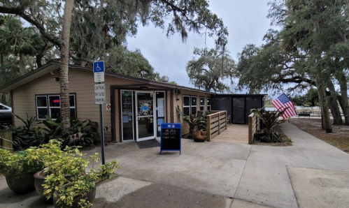 A small building with a wooden deck, surrounded by greenery, featuring an American flag and a sign for a restaurant.