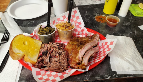 A red plate with pulled pork, ribs, collard greens, coleslaw, and a slice of cornbread on a checkered tablecloth.