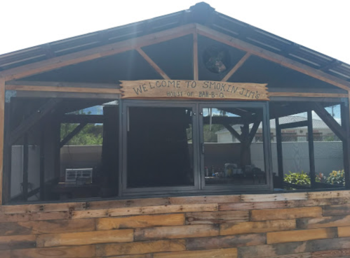 A wooden structure with a sign reading "Welcome to Smokin J's House of Bar-B-Q," featuring large windows and a rustic design.
