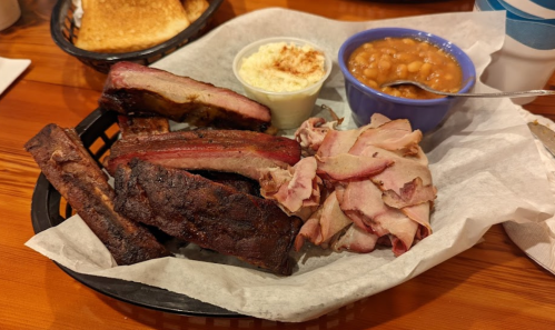 A basket of barbecue ribs, sliced meat, baked beans, and a side of creamy coleslaw on a wooden table.