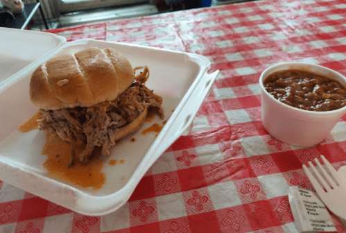 A pulled pork sandwich on a bun with barbecue sauce, next to a cup of baked beans on a red-checkered tablecloth.