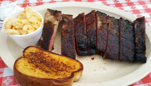 A plate with sliced smoked brisket, a side of macaroni and cheese, and a piece of toasted bread on a checkered tablecloth.