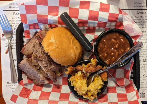 A plate with a pulled pork sandwich, baked beans, and cornbread, served in a red and white checkered basket.