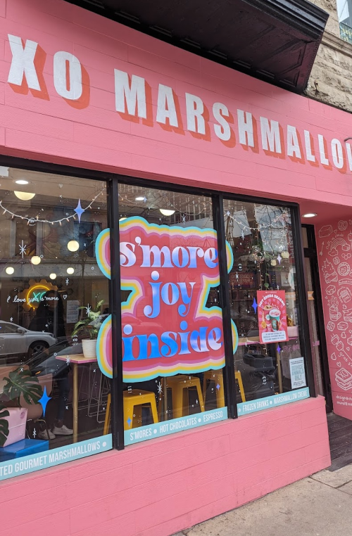 A colorful storefront with "s'more joy inside" sign, showcasing gourmet marshmallows and cozy seating.