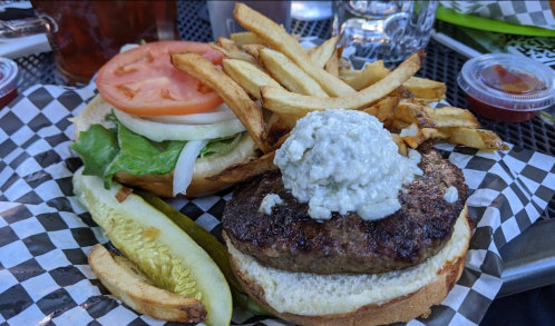 A burger with lettuce, tomato, and onion, topped with a creamy sauce, served with fries and a pickle on a checkered plate.