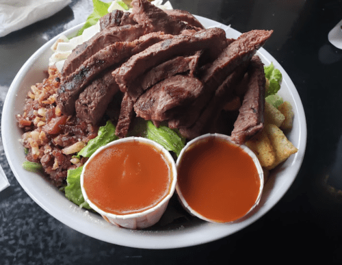 A bowl filled with sliced steak, lettuce, rice, and two cups of sauce on the side.