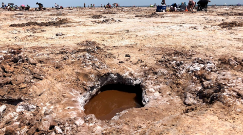 A large, muddy hole surrounded by salt flats, with people enjoying the beach in the background.