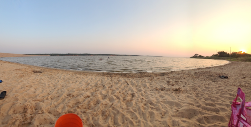 A serene beach scene at sunset, with gentle waves lapping at the sandy shore and a few people in the water.