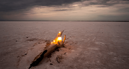 A weathered log with candles sits on a vast, salt flat under a moody, overcast sky at dusk.