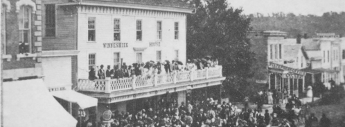 Historic black-and-white photo of a large crowd outside a hotel, with people on the balcony and buildings in the background.