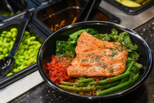A bowl with salmon on a bed of greens, topped with green beans and red sauce, alongside edamame in the background.