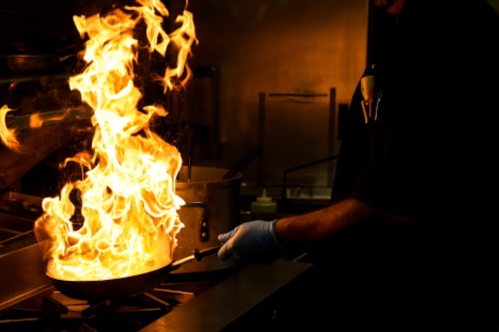 A chef in a dark kitchen ignites a pan, creating a dramatic burst of flames.