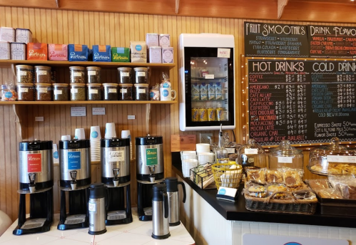 A cozy café counter with coffee machines, snacks, and a variety of drink options displayed on shelves.