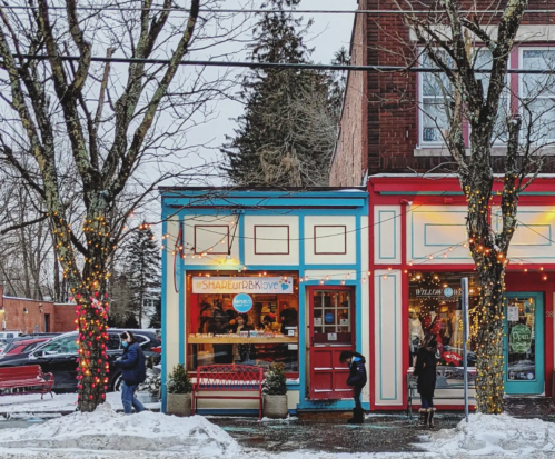Colorful storefronts adorned with holiday lights, surrounded by snow and trees, with people walking by.
