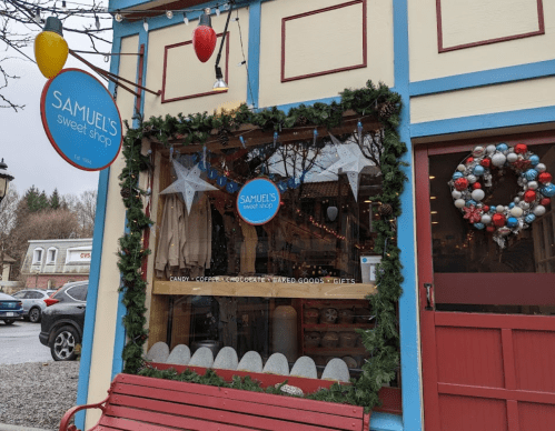 A charming storefront of Samuel's Sweet Shop, decorated with holiday lights and greenery, featuring a festive wreath.